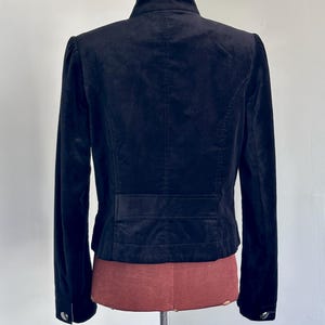 May include: A black velvet jacket with a high collar and long sleeves. The back of the jacket features a tailored design with a horizontal seam detail at the waist and buttoned cuffs. The jacket is displayed on a mannequin.