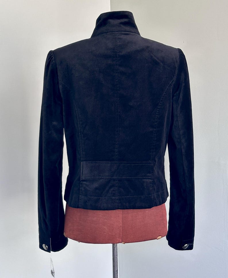 May include: A black velvet jacket with a high collar and long sleeves. The back of the jacket features a tailored design with a horizontal seam detail at the waist and buttoned cuffs. The jacket is displayed on a mannequin.