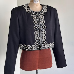 May include: A black jacket with long sleeves and a decorative beaded design. The jacket features a round neckline and a cropped length. The embellishments include silver and white beads arranged in geometric patterns.