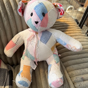 May include: A handmade patchwork teddy bear with a variety of pastel-colored fabric patches. The bear has black button eyes, a stitched nose, and floral fabric on the ears. A pink ribbon is tied around its neck. The bear is sitting on a textured, light brown surface.