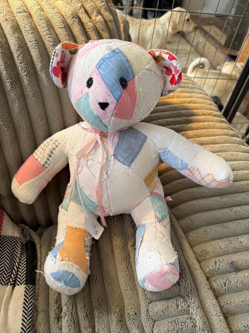 May include: A handmade patchwork teddy bear with a variety of pastel-colored fabric patches. The bear has black button eyes, a stitched nose, and floral fabric on the ears. A pink ribbon is tied around its neck. The bear is sitting on a textured, light brown surface.