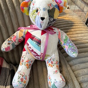 May include: A handmade patchwork teddy bear with a variety of floral and patterned fabrics. The bear has a pink ribbon around its neck and features colorful ears. The bear is sitting on a textured surface.