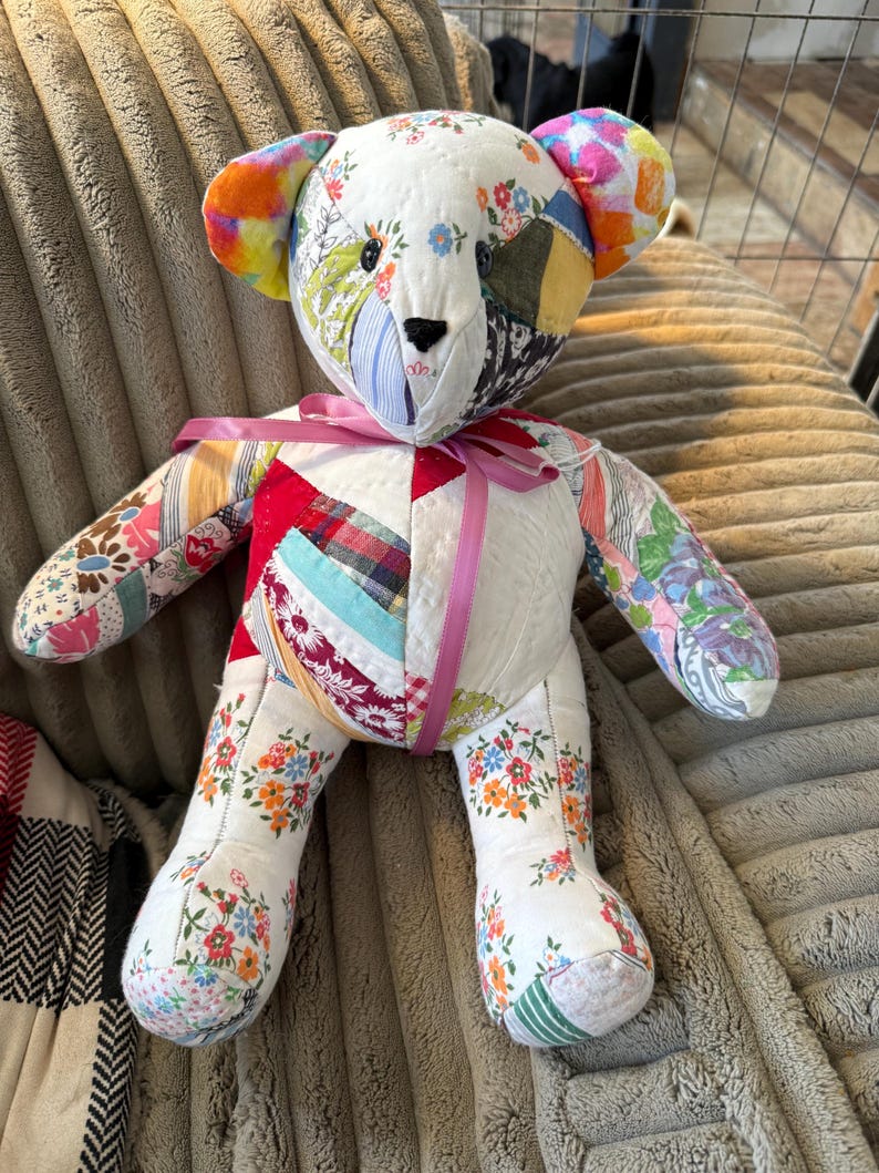 May include: A handmade patchwork teddy bear with a variety of floral and patterned fabrics. The bear has a pink ribbon around its neck and features colorful ears. The bear is sitting on a textured surface.