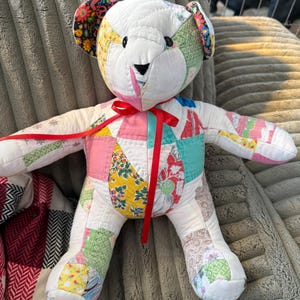 May include: A handmade patchwork teddy bear with a white body and colorful fabric patches. The bear has a red ribbon tied around its neck and features black button eyes and a black stitched nose. The ears are adorned with floral fabric.