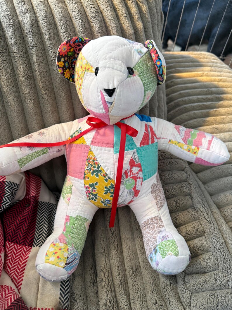 May include: A handmade patchwork teddy bear with a white body and colorful fabric patches. The bear has a red ribbon tied around its neck and features black button eyes and a black stitched nose. The ears are adorned with floral fabric.