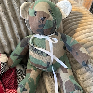 May include: A handmade teddy bear crafted from camouflage fabric. The bear features a white ribbon tied around its neck and the name "GOWER" stitched on its chest. The bear has button eyes and a stitched nose.