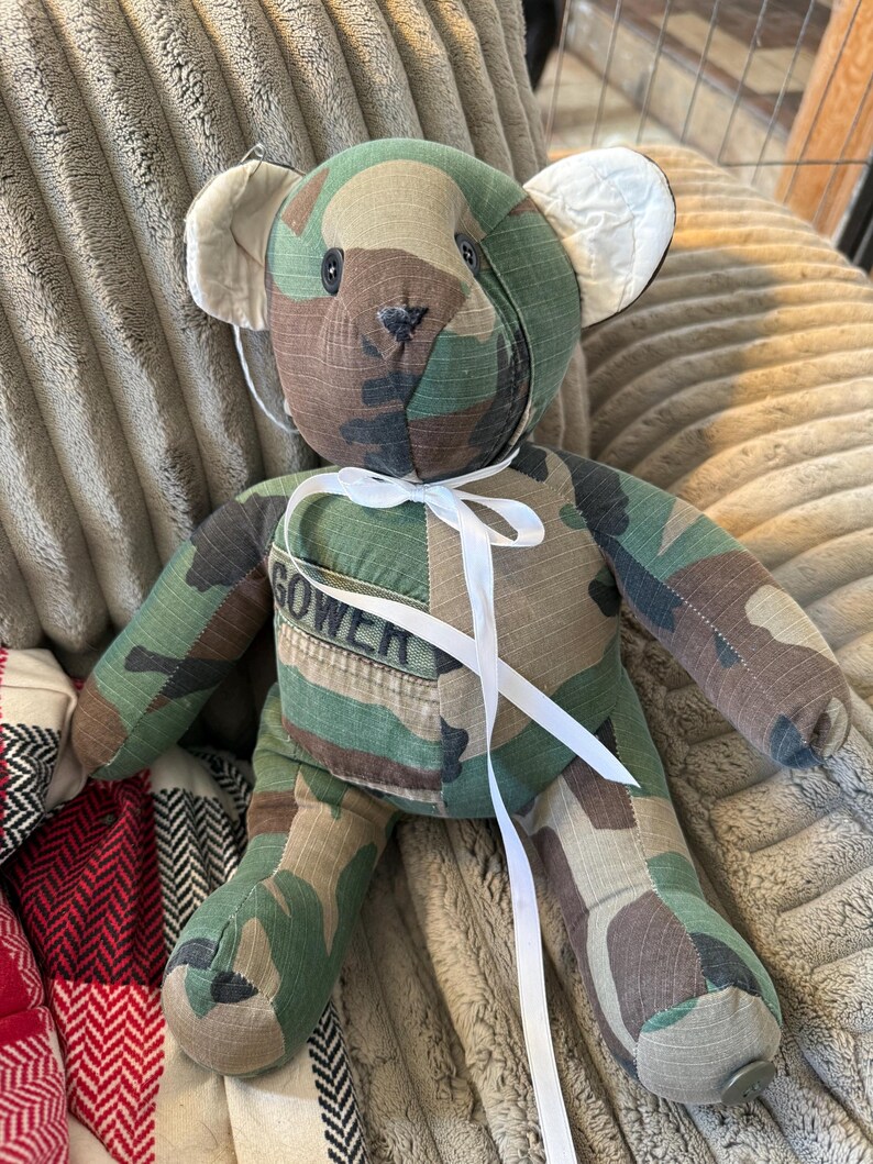 May include: A handmade teddy bear crafted from camouflage fabric. The bear features a white ribbon tied around its neck and the name "GOWER" stitched on its chest. The bear has button eyes and a stitched nose.