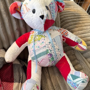 May include: Handmade patchwork teddy bear with a white head and limbs, red arms and legs, and a body adorned with colorful fabric patches. The bear has black button eyes, a black nose, and a light blue ribbon tied around its neck. A unique, handcrafted toy.