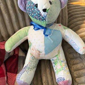 May include: A handmade patchwork teddy bear with a variety of floral and patterned fabrics in pastel colors. The bear has a blue ribbon around its neck and black button eyes. The ears are lavender.