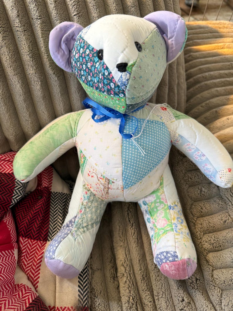 May include: A handmade patchwork teddy bear with a variety of floral and patterned fabrics in pastel colors. The bear has a blue ribbon around its neck and black button eyes. The ears are lavender.