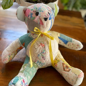 May include: A patchwork teddy bear with a yellow ribbon around its neck. The bear is made of various fabrics in shades of pink, blue, green, and white.
