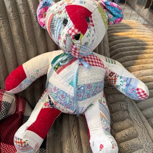 May include: Handmade patchwork teddy bear with a variety of fabric patterns, including floral, plaid, and striped designs. The bear has black button eyes, red accents, and a light blue ribbon tied around its neck. A unique, handcrafted item.
