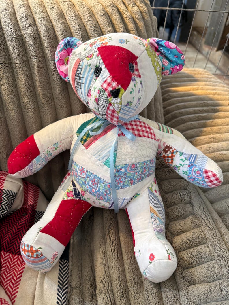 May include: Handmade patchwork teddy bear with a variety of fabric patterns, including floral, plaid, and striped designs. The bear has black button eyes, red accents, and a light blue ribbon tied around its neck. A unique, handcrafted item.