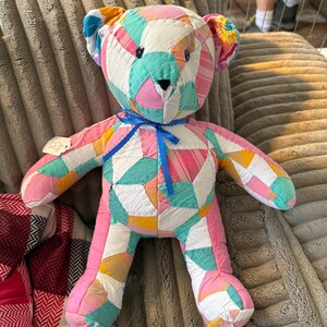 May include: A handmade patchwork teddy bear in a variety of colors including pink, blue, yellow, and white. The bear has a blue ribbon around its neck and is made of fabric pieces sewn together. A tag is attached to the bear's left arm.
