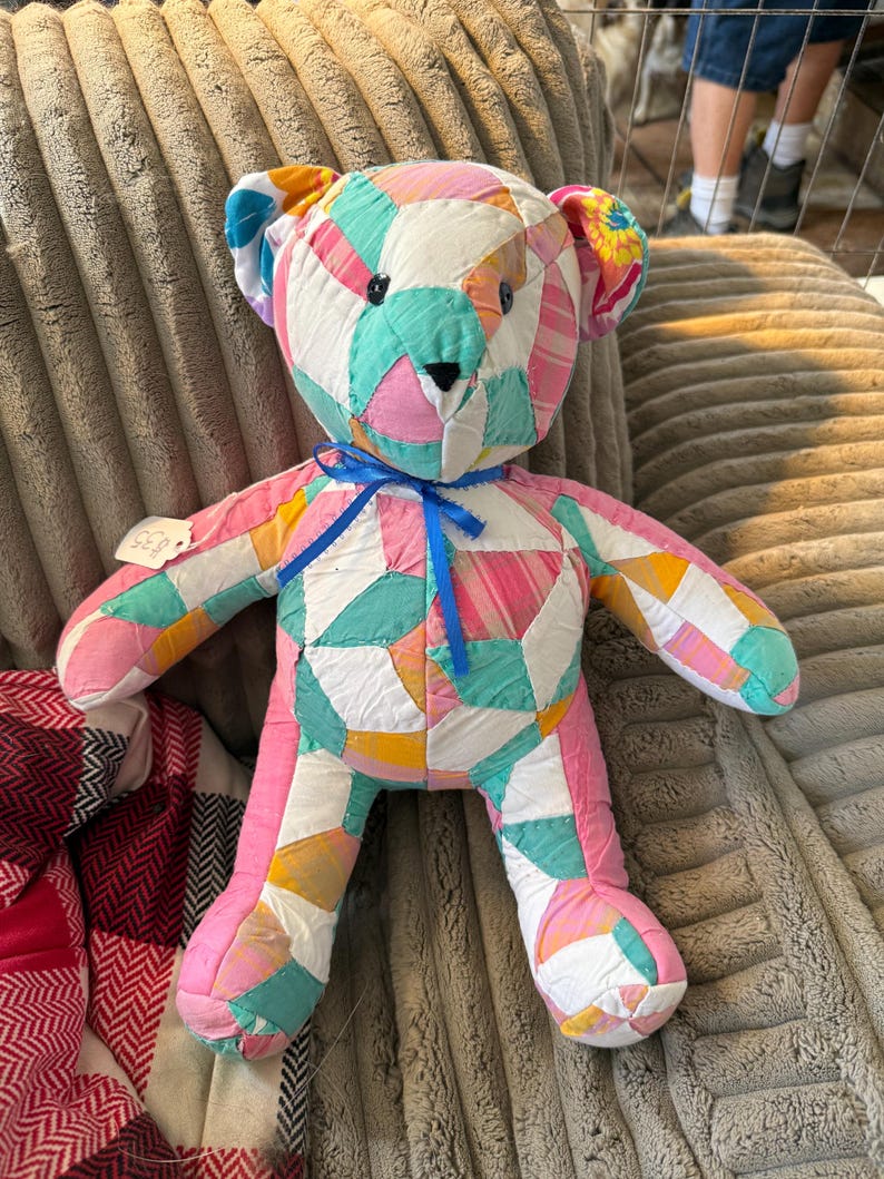 May include: A handmade patchwork teddy bear in a variety of colors including pink, blue, yellow, and white. The bear has a blue ribbon around its neck and is made of fabric pieces sewn together. A tag is attached to the bear's left arm.