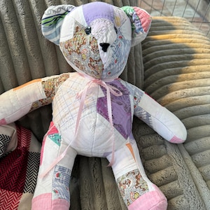 May include: A handmade patchwork teddy bear with a variety of fabric patterns and colors. The bear has a pink ribbon tied around its neck and pink fabric on the soles of its feet. The bear is sitting on a gray, textured surface.