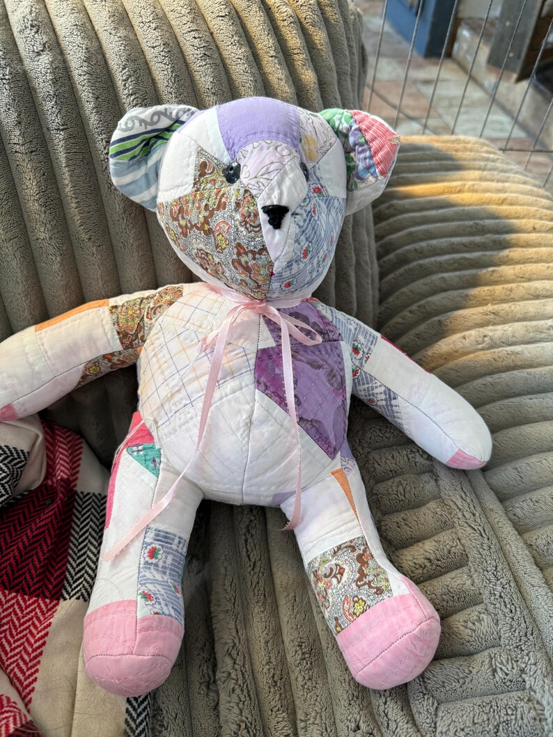 May include: A handmade patchwork teddy bear with a variety of fabric patterns and colors. The bear has a pink ribbon tied around its neck and pink fabric on the soles of its feet. The bear is sitting on a gray, textured surface.