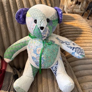 May include: A handmade patchwork teddy bear with a variety of fabric patterns in shades of white, green, blue, and purple. The bear has black button eyes and a green ribbon tied around its neck. The bear is sitting on a textured surface.