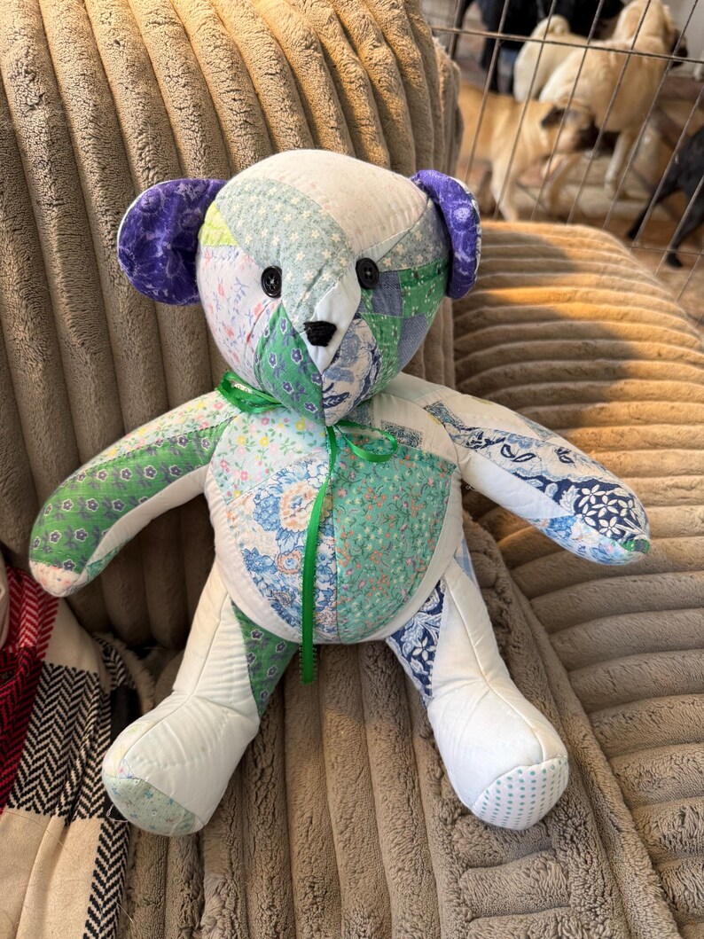 May include: A handmade patchwork teddy bear with a variety of fabric patterns in shades of white, green, blue, and purple. The bear has black button eyes and a green ribbon tied around its neck. The bear is sitting on a textured surface.