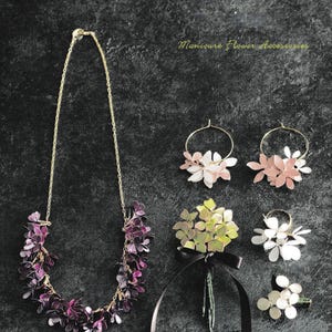 Peut inclure: Un ensemble d'accessoires de bijoux en forme de fleurs, comprenant un collier, des boucles d'oreilles, une bague et une broche. Le collier est fait de fleurs violettes, les boucles d'oreilles sont roses, la bague est blanche et la broche est verte. Les bijoux sont présentés sur un fond gris foncé.