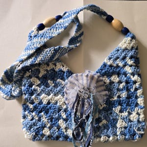 May include: A handmade crochet bag in shades of blue and white, featuring a floral embellishment and a striped strap. The strap is adorned with wooden beads. The bag's open weave design is visible. The bag is approximately 12 inches wide.