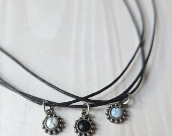 May include: A multi-strand black cord necklace featuring three small circular pendants. Each pendant has a silver-toned scalloped edge and a central stone: one white with grey veining, one solid black, and one light blue with white veining.