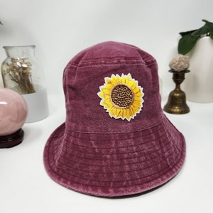 May include: A burgundy bucket hat with a yellow and brown sunflower patch. The hat has a soft, textured appearance and a wide brim. The sunflower patch is centered on the front of the hat.