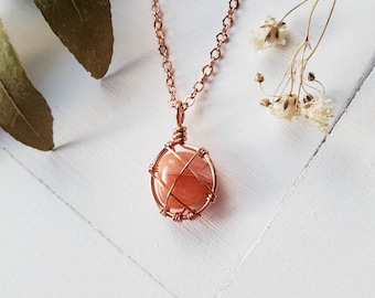 May include: A copper-colored necklace with a pendant featuring an orange and white stone encased in a wire cage. The chain and pendant are set against a white wooden surface, with dried flowers and leaves in the background.