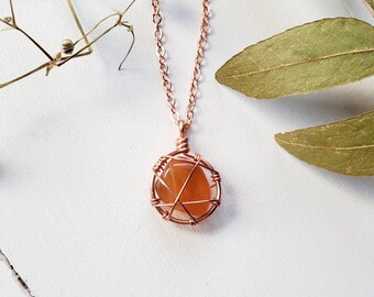 May include: A copper-colored wire-wrapped pendant necklace featuring a round, orange gemstone. The pendant hangs from a delicate chain. The necklace is displayed on a white surface with dried flowers and leaves.