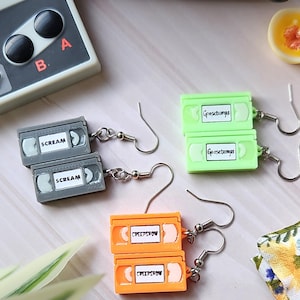 May include: A set of four pairs of earrings shaped like VHS tapes. Each pair features a different movie title: "Scream", "Goosebumps", and "Creepshow". The earrings are made of plastic and have silver hooks.