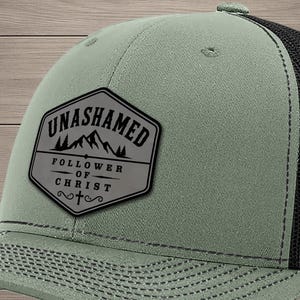 May include: A loden green baseball cap with a black mesh back and a structured front panel. A hexagonal patch on the front features the text UNASHAMED FOLLOWER OF CHRIST above a mountain graphic and a small cross symbol in black ink.