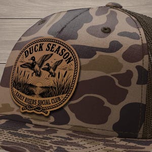 May include: A camouflage baseball cap with a brown mesh back and a circular rawhide patch on the front. The patch features an illustration of two ducks flying over a marsh with the text DUCK SEASON and EARLY RISERS SOCIAL CLUB engraved.