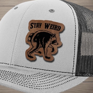 May include: A heather gray and black trucker hat featuring a tan leather-like patch on the front. The patch displays a black illustration of a raccoon and the text STAY WEIRD in a bold, dark font. The hat has a structured crown and mesh back.