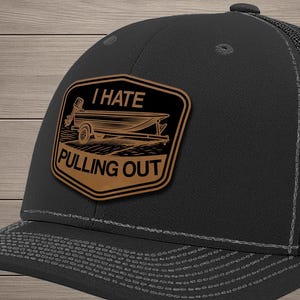 May include: A black trucker hat featuring a brown leather-like patch on the front. The patch displays a graphic of a boat on a trailer with the text I HATE PULLING OUT. The hat has a solid front panel and a black mesh back with a curved brim.