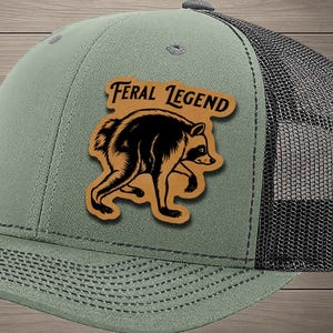 May include: An olive green trucker hat with a black mesh back and a tan leather patch on the front. The patch features a black graphic illustration of a raccoon and the text Feral Legend printed in a bold, dark font above the animal figure.