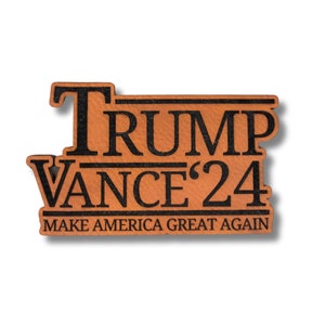 May include: A wooden pin with the words "TRUMP VANCE '24" in black lettering. Below the names, the text "MAKE AMERICA GREAT AGAIN" is also in black. The pin has a light brown background and a shadow.