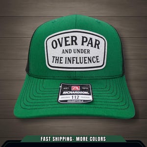 Pode incluir: Boné de caminhoneiro verde com parte traseira em malha preta e um patch branco. O patch diz "OVER PAR AND UNDER THE INFLUENCE". Uma pequena etiqueta abaixo diz "RICHARDSON ORIGINAL 112 TRUCKER ADJUSTABLE".