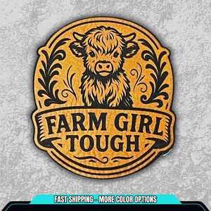 May include: A round, wooden-style emblem with a Highland cow illustration. The emblem features the text "FARM GIRL TOUGH" in a banner across the bottom. The cow is in the center, with black decorative flourishes around the edges.