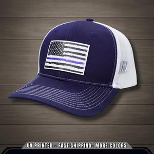 Pode incluir: Boné de caminhoneiro azul marinho e branco com um patch com um design de bandeira americana desgastada e uma linha azul fina. O boné tem uma parte traseira de malha e uma aba curva. Costuras brancas na aba e na coroa.