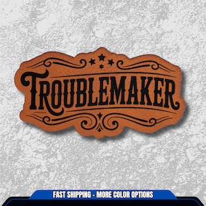 May include: A brown leather patch with the word "TROUBLEMAKER" in bold black lettering. The patch has decorative swirls and stars, with a vintage, western-inspired design. The patch is likely for clothing or accessories.
