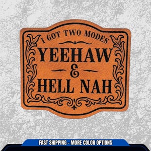 May include: A rectangular, brown leather patch with black text and decorative flourishes. The text reads "I GOT TWO MODES YEEHAW & HELL NAH". The patch has a curved top and bottom edge.