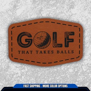 May include: A brown leather patch with the word "GOLF" and a golf ball graphic in black. Below the word "GOLF" is the phrase "THAT TAKES BALLS" in black. The patch has a stitched border and is set against a textured gray background.