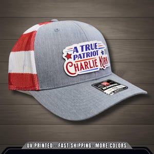 Op de afbeelding: Een grijze baseballcap met een rood en wit mesh achterpaneel. De cap heeft een patch met de tekst "A True Patriot Charlie Kirk" in rood, wit en blauw. De cap heeft een gebogen klep en een Richardson 112 label.