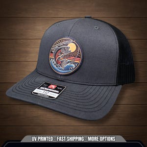 May include: A dark grey baseball cap with a black mesh back. The cap has a circular patch with a colourful illustration of a fish leaping from water, with a sun behind. The Richardson 112 adjustable hat has a tag.