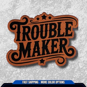 May include: A brown leather-like patch with the words "TROUBLE MAKER" in bold black letters. The design includes decorative swirls, stars, and a banner. The patch has a vintage, rustic aesthetic.