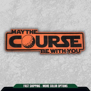 May include: An orange rectangular sign with the text "MAY THE COURSE BE WITH YOU" in black. A golf ball graphic is in the center. The sign is on a textured gray background. A fun gift for golfers.