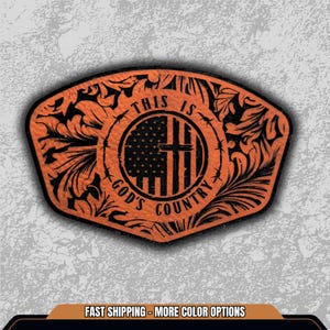 May include: An orange patch with black filigree and the text "THIS IS GOD'S COUNTRY." The design includes an American flag with a cross, surrounded by barbed wire. The patch has a black border.