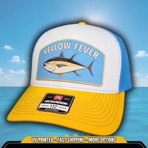May include: A white and blue trucker hat with a yellow bill. The front of the hat features a patch with a yellowfin tuna and the text "YELLOW FEVER". The hat also has a Richardson 112 Trucker label.