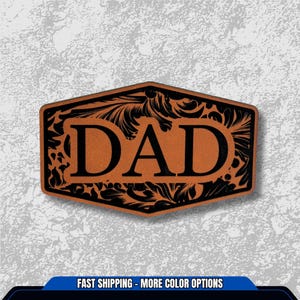 May include: A brown leather patch with the word "DAD" in bold black letters. The patch has a decorative black floral design around the edges. The patch is rectangular with a slightly curved top and bottom.