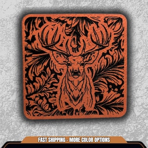 May include: Square coaster with a deer head design. The coaster features a detailed black illustration of a stag against an orange background. The deer is centered, surrounded by a floral pattern.